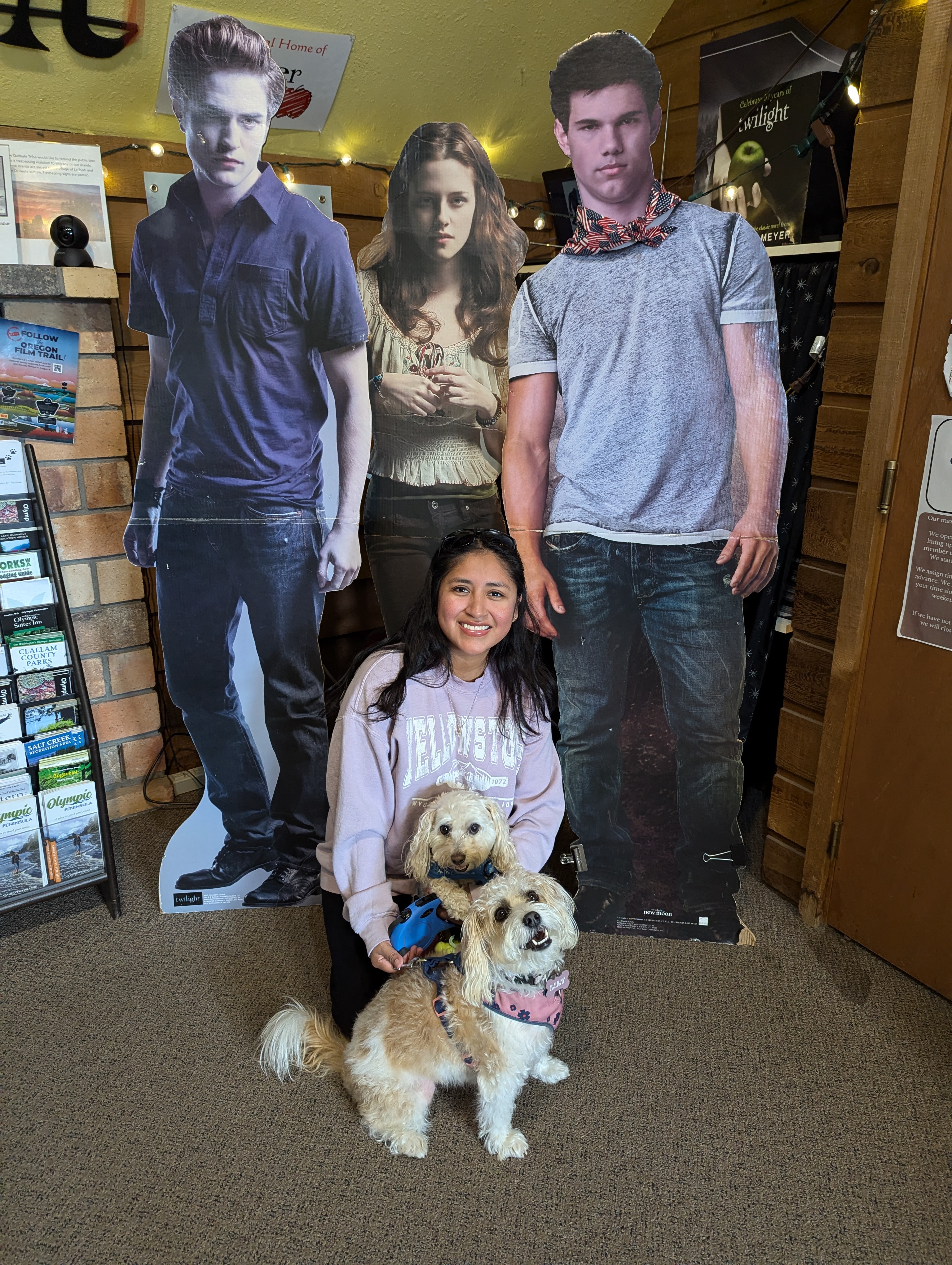 Minerva Barrios, a petite woman with black hair, kneels indoors smiling between two small, fluffy dogs while posing in front of life-size cardboard cutouts of characters from the Twilight series in Forks, Washington