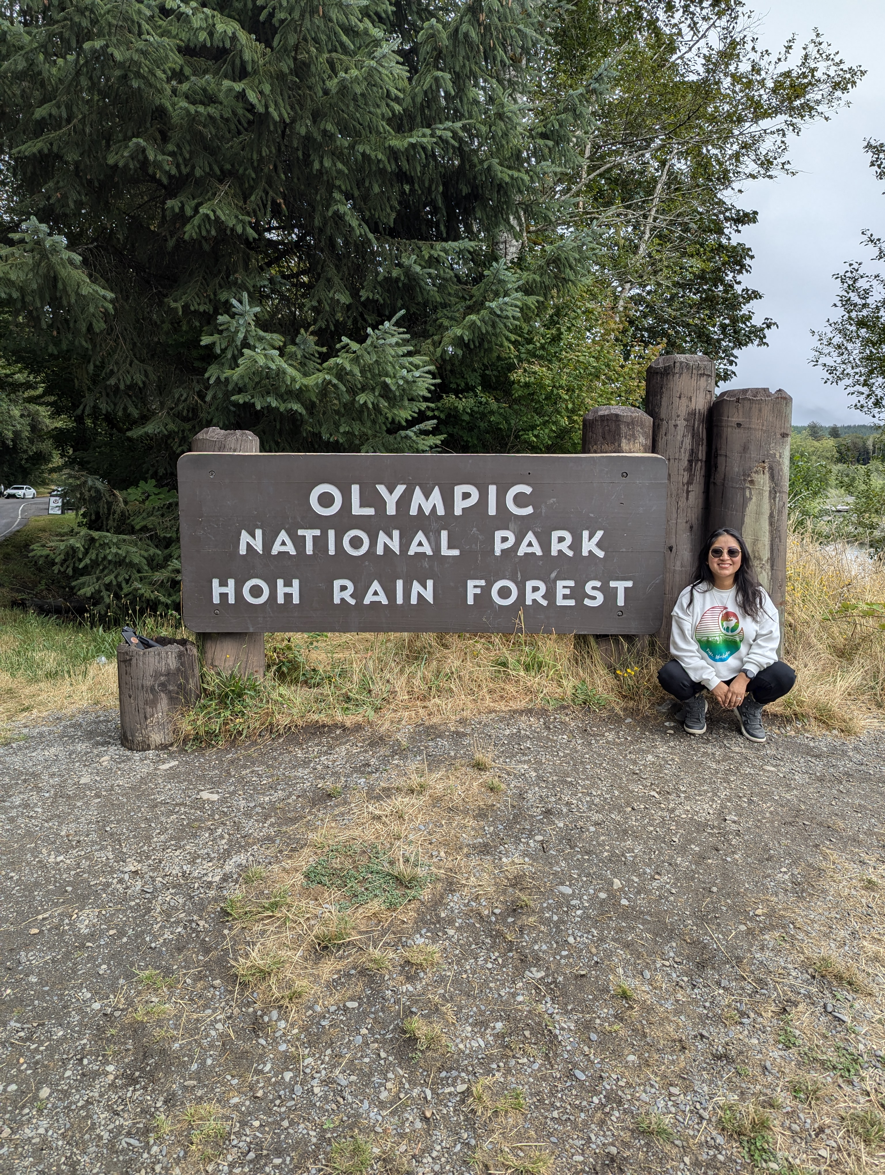 Minerva Barrios, a petite woman with black hair and sunglasses, crouches beside a large wooden sign that reads “Olympic National Park – Hoh Rain Forest,” with lush green trees in the background