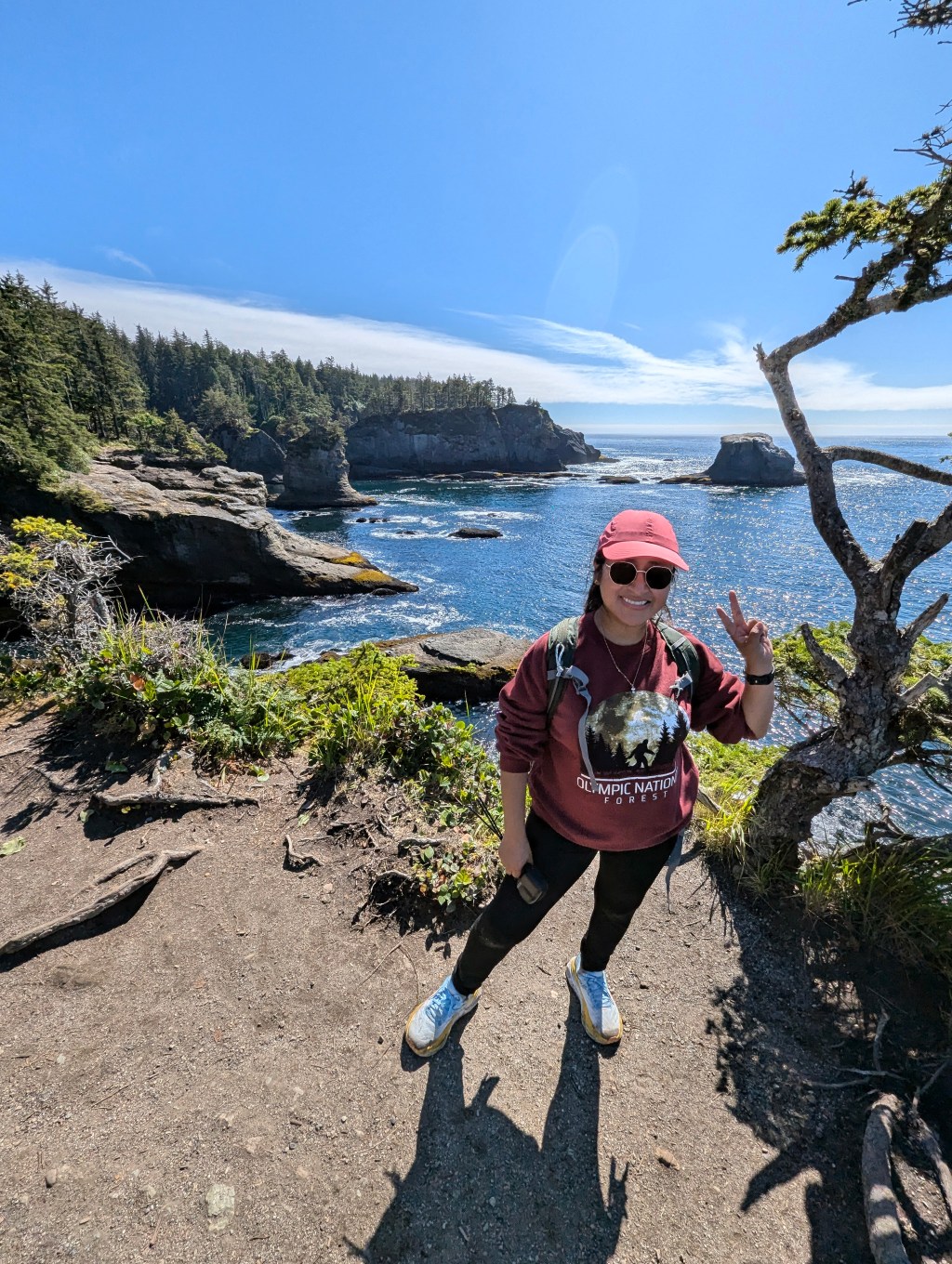 Minerva Barrios, a petite woman with straight black hair, stands at the edge of Cape Flattery, Washington, with rugged coastal cliffs and the Pacific Ocean in the background. She faces the camera, smiling and holding up a peace sign with her right hand. Overcast skies and windswept evergreen trees frame the scenic, windswept landscape