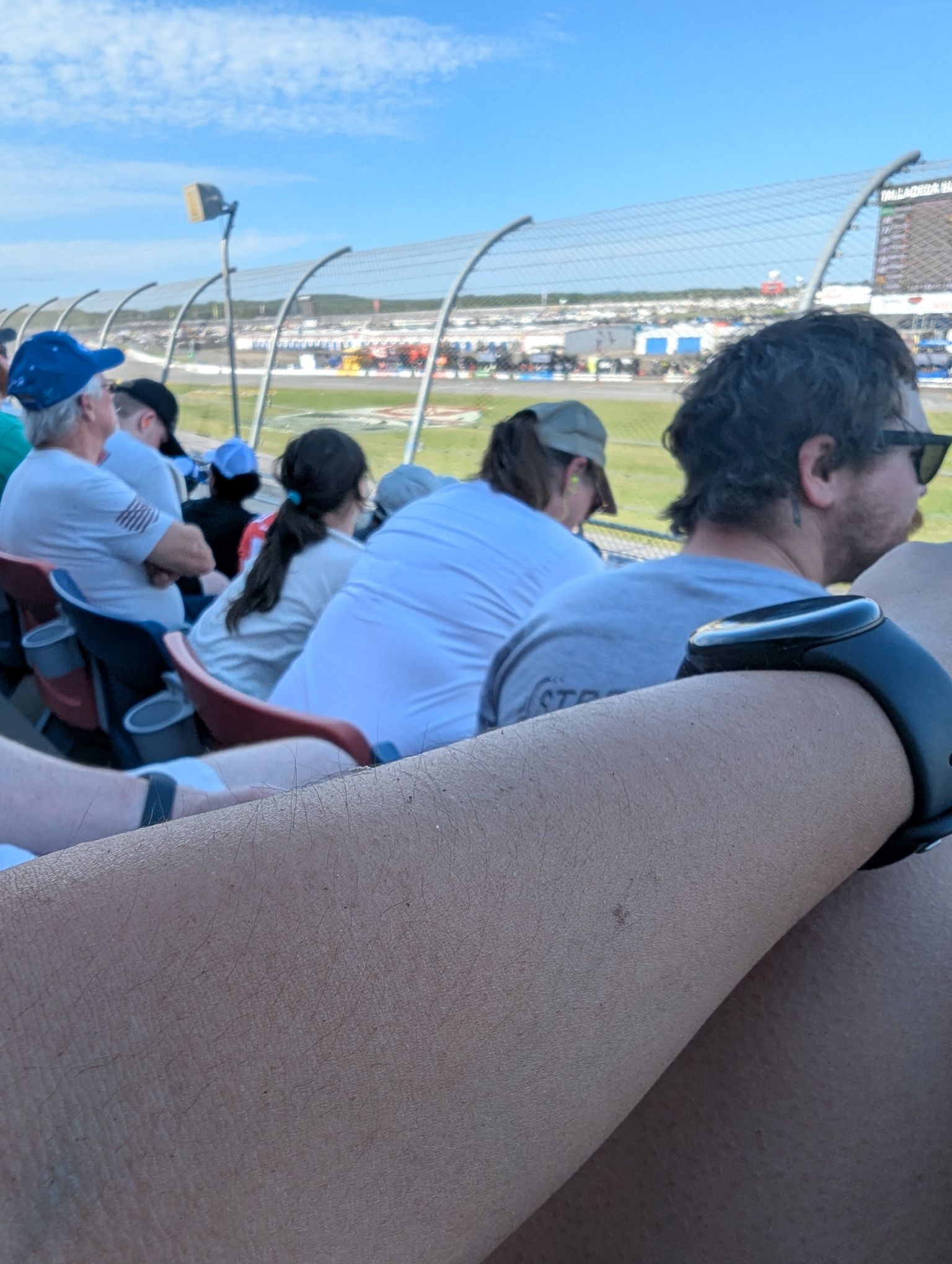 Fans seated in a packed stadium overlook a racetrack through a chain-link fence, with the large scoreboard at Talladega Superspeedway visible in the background. A person’s forearm wearing a smartwatch is prominently in the foreground